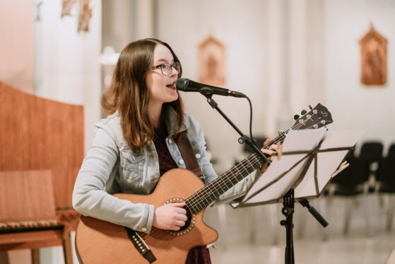 Hannah Stienen steht in der Kirche mit Gitarre, Mikrofon und Notenständer und singt moderne Tauflieder.