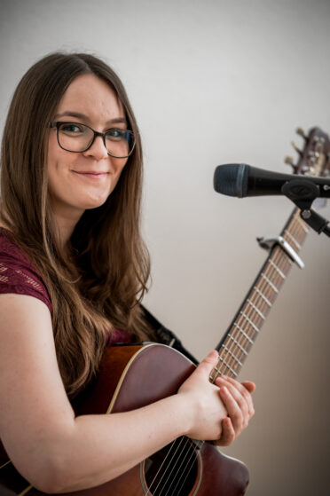 Sängerin Hannah Stienen steht mit ihrer Gitarre am Mikrofon bei einem Firmenevent und schaut in die Kamera.
