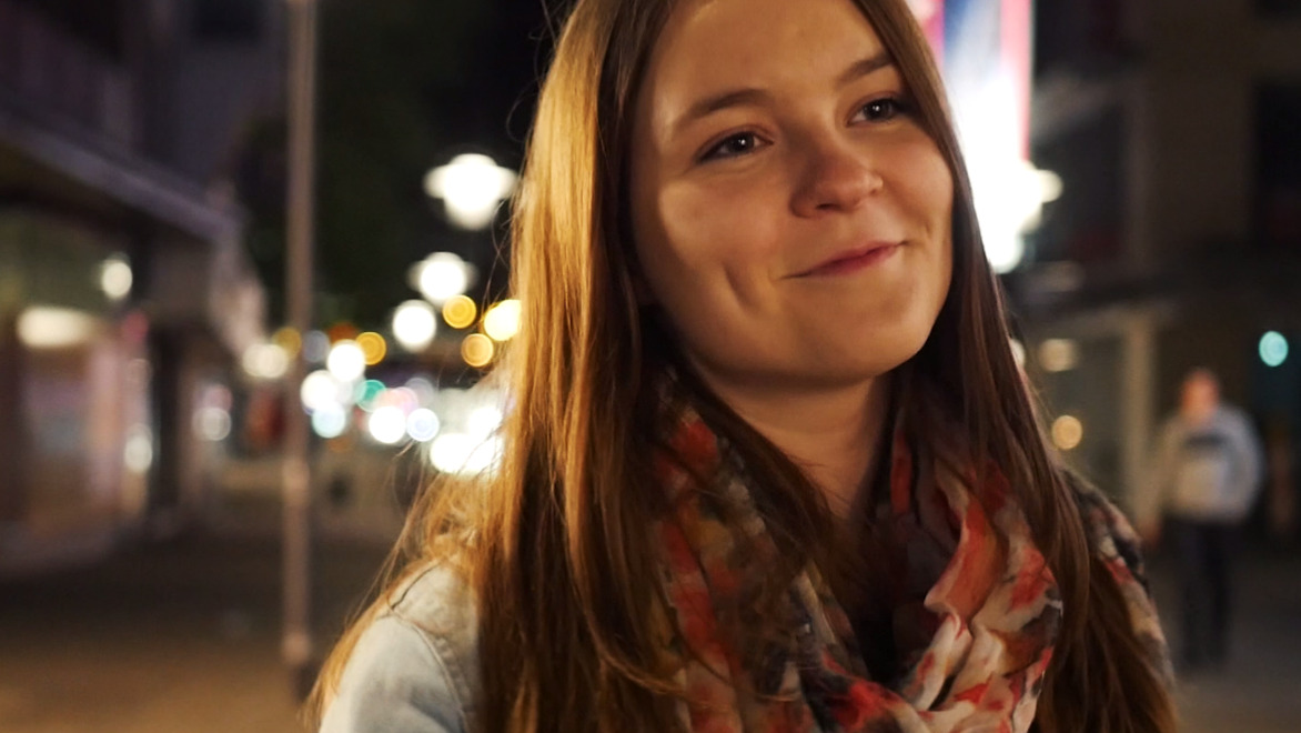Dieses Bild zeigt Hannah im Profil. Es ist ein Standbild aus dem Musikvideo für den Song "Wenn Sie Lacht", wie sie lächelt und in der nächtlichen Stadt spazieren geht. Die Lichter leuchten unscharf im Hintergrund.