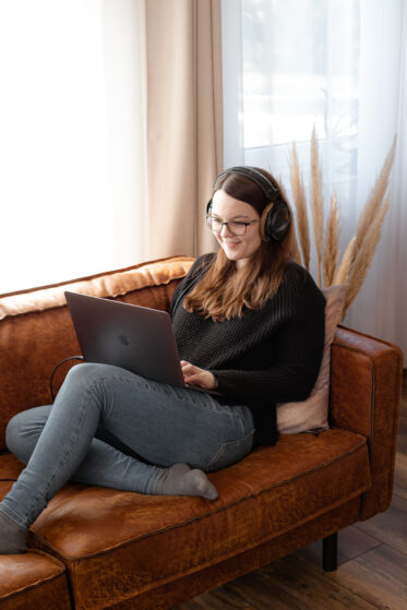 Hannah Stienen sitzt auf einer Couch mit ihrem Laptop und hat Kopfhörer auf. Sie arbeitet an einem persönlichen Song für ein Kundenpaar.