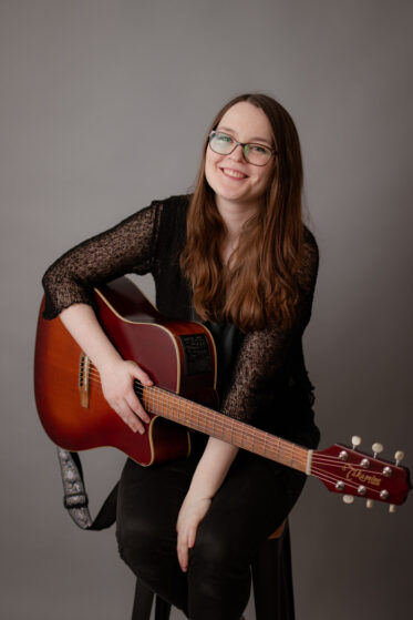 Taufsängerin Hannah Stienen sitzt mit ihrer Gitarre auf einem Hocker vor einer grauen Wand und lacht in die Kamera.