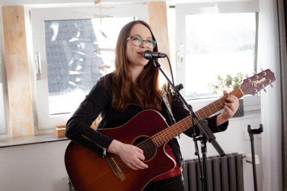 Hannah Stienen bei einem Wohnzimmerkonzert. Sie steht mit Gitarre und Mikrofon vor einer Fensterwand und singt.