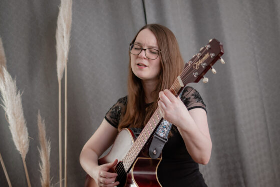 Hannah Stienen steht als Sängerin zur Beerdigung mit ihrer Gitarre vor einem grauen Vorhang und singt.