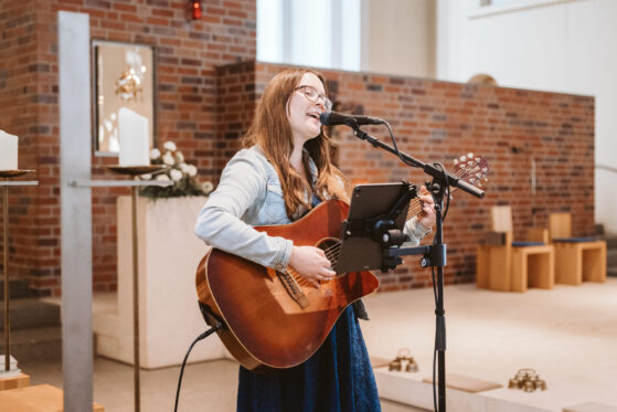 Moderne Tauflieder (c) Jörg Brücker Hannah Stienen steht in der Kirche mit Gitarre, Mikrofon und Notenständer und singt moderne Tauflieder.